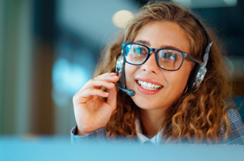 Mujer con cabello rizado y auriculares sonríe durante una llamada. Entorno profesional de atención al cliente.